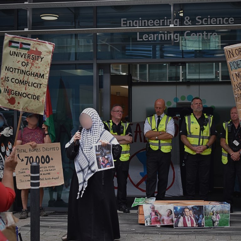 protesters holding signs, a woman addressing them and police in the background