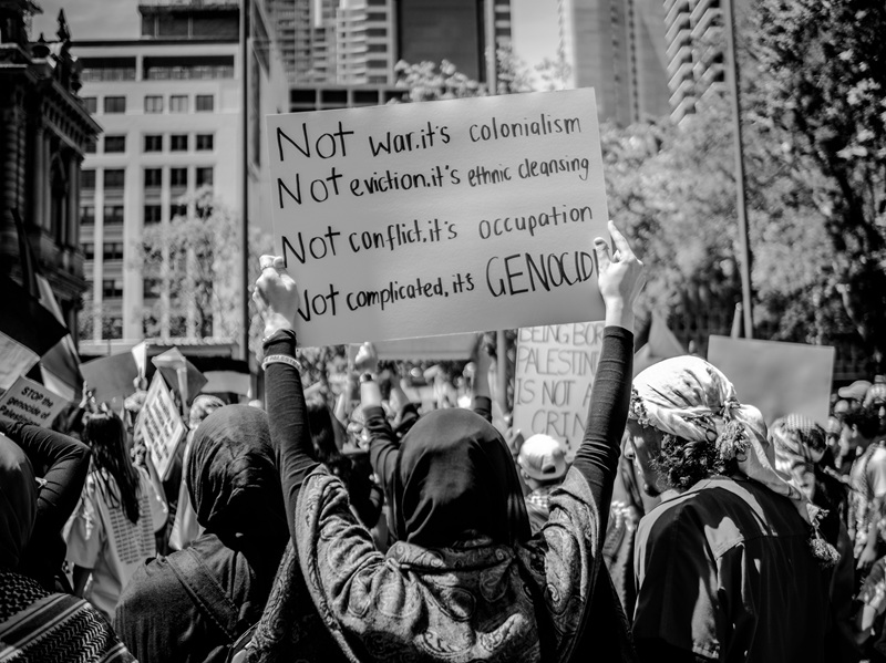 black and white image showing a woman holding a poster in a rally: Not complicated, It is Genocide